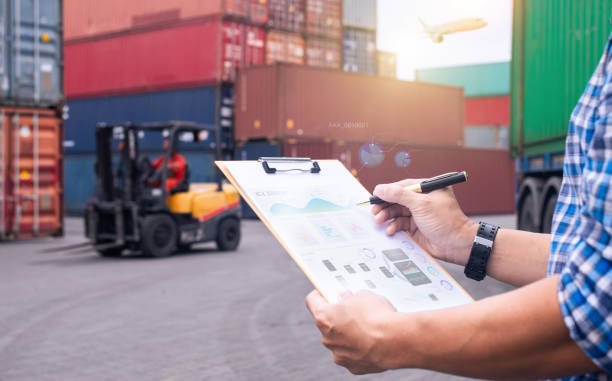 Logistics manager reviewing documents at a shipping yard, with a forklift and cargo containers in the background.